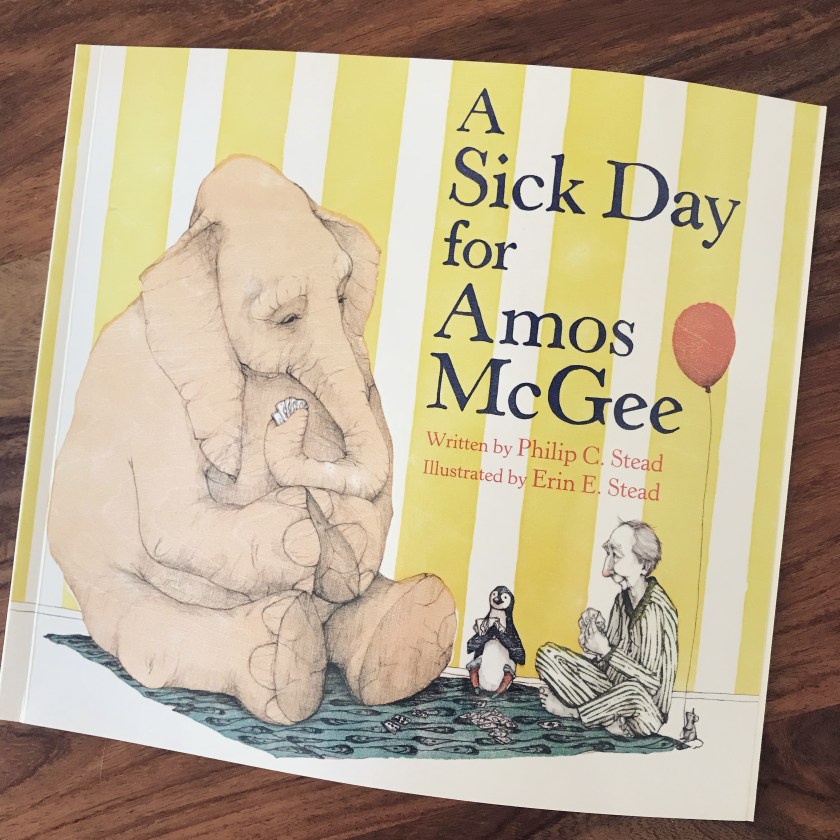 The image is a photograph of the cover of A Sick Day for Amos McGee against a wooden table background. The cover is a drawing of an elephant, a penguin, and a man playing cards. They are sitting on a blue patterned blanket and there is a small mouse holding a large red balloon next to the man.