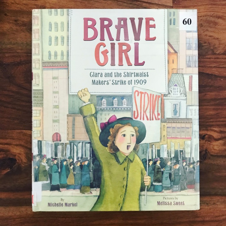 A photo of a book cover. The cover says "Brave Girl: Clara and the Shirtwaist Makers' Strike of 1909" and features a drawing of a woman dressed in a green coat with one fist in the air and holding a picket sign that says "strike." In the background are women marching through the city and holding picket signs. 